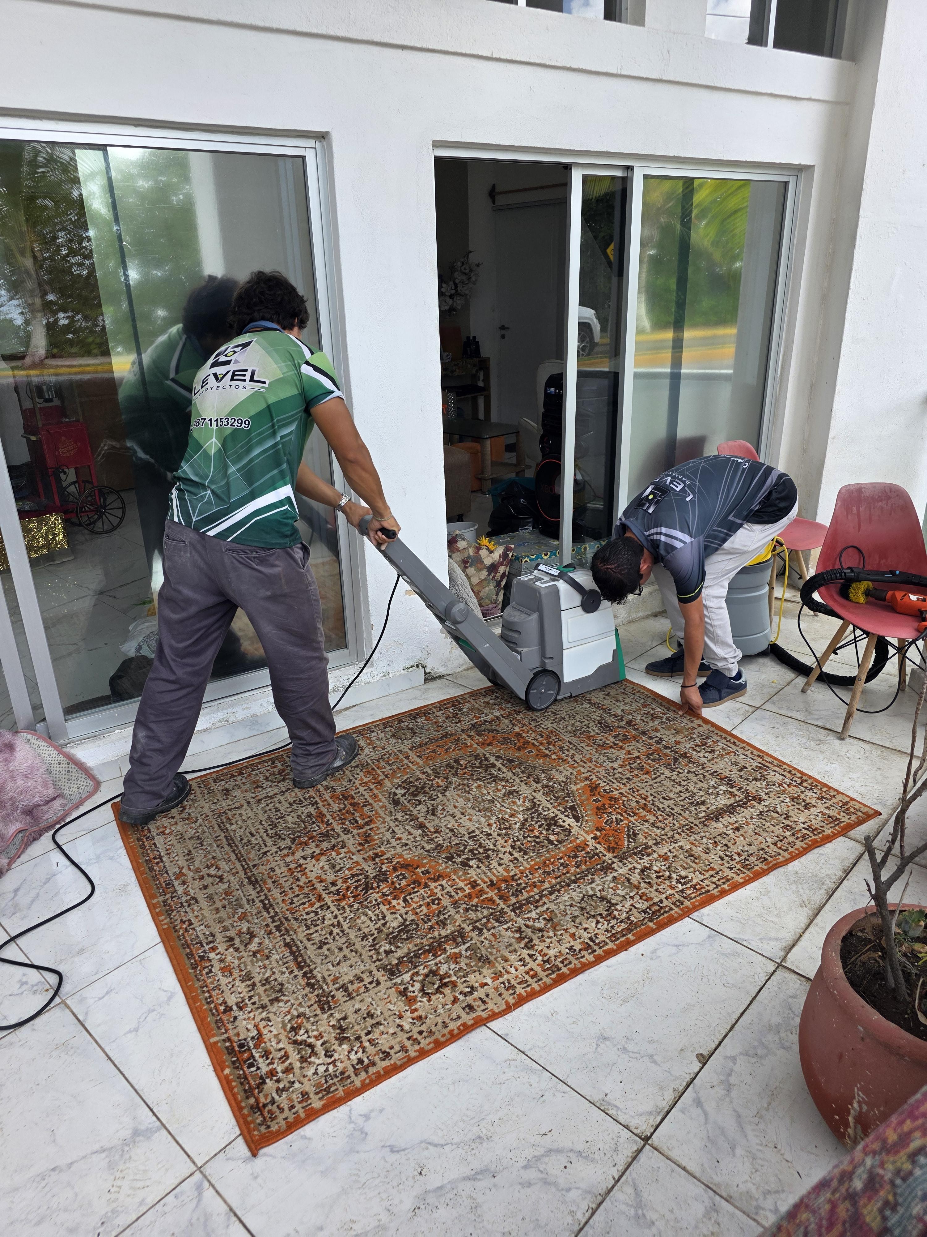Dos personas limpiando una alfombra con una máquina cerca de puertas corredizas de vidrio.