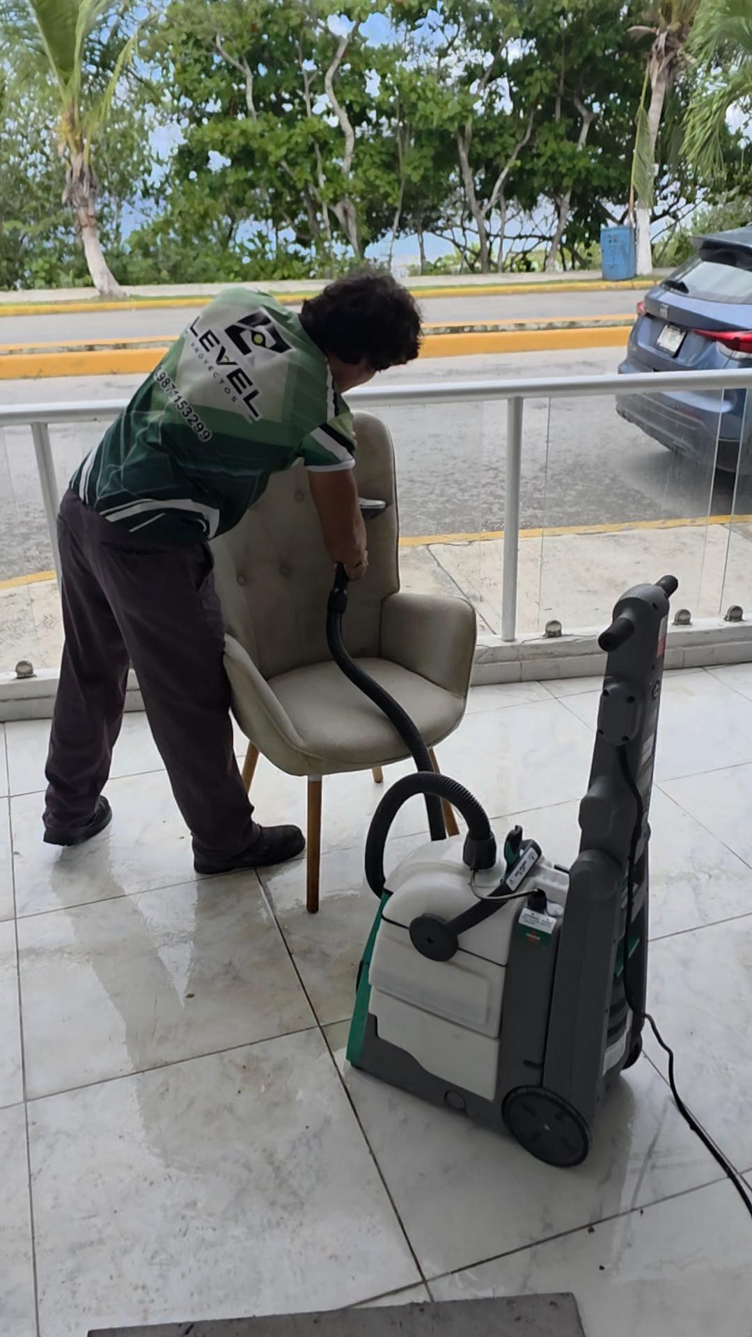 Persona usando una aspiradora para limpiar una silla tapizada en un área con ventanales.